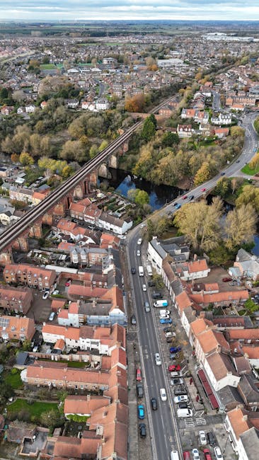 An aerial view of a residential area in Motspur Park showing a mix of terraced and semi-detached houses with red-brick exteriors and tiled roofs. A prominent road runs through the centre of the image, with numerous cars parked along the sides and some moving along the street. Adjacent to the road, there is a line of closely packed houses with small gardens and driveways, some with visible pathways and outdoor furniture. To the left, a railway line runs parallel to the road, with a railway bridge crossing over the road and a stretch of trees and green spaces beyond. The background features a large park or wooded area with dense foliage and open fields stretching into the distance, under an expansive sky with scattered clouds. This scene highlights a typical neighbourhood within the Motspur Park area, which [COMPANY_NAME] can assist with in home relocation and furniture transport services, involving careful packing and loading processes.