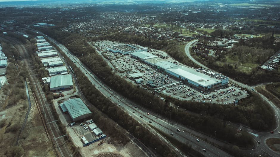 An aerial view showing a large commercial building with a substantial parking lot, surrounded by trees and open land, situated adjacent to a busy road with multiple lanes of traffic. The parking area contains numerous cars, and the building appears to be part of a warehouse or retail complex. Nearby, there are smaller structures, including a warehouse with a curved roof and a few other industrial-type buildings. The scene is illuminated by natural daylight, with clear visibility of the surrounding landscape. This image captures the environment typical of a commercial area involved in logistics, potentially supporting home or office relocations by facilitating furniture transport and packing during moves, as observed through the large-scale premises and vehicle access routes related to professional removals, such as those provided by manwithvanmotspurpark.co.uk.