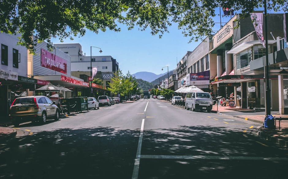 A wide view of a commercial street in Motspur Park on a clear day, showing storefronts with signage and awnings, parked cars along both sides including a silver and a white van, and pedestrians walking on the sidewalks. Trees with green foliage cast shadows on the pavement, and in the background, a mountain range is visible beneath a blue sky with minimal clouds. The scene captures the lively atmosphere of shopping and small business activity typical of a local shopping district, with some shop displays and outdoor seating areas present. This environment, associated with residential and commercial properties, often features in local home and business relocations, with trucks and relocation furniture possibly occupying the street during moving processes, as seen with the presence of vehicles and activity consistent with moving logistics or packing and furniture transport.