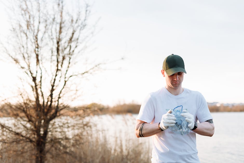 A man wearing a dark baseball cap, white T-shirt, and white gloves is standing outdoors near a body of water, with a large, leafless tree in the background during daytime. He appears to be inspecting