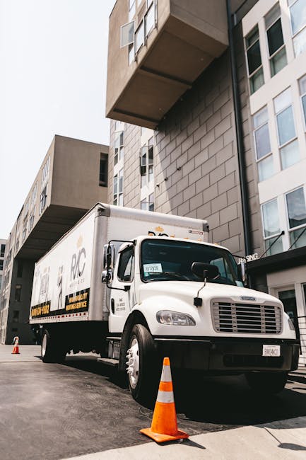 A white box truck with black and yellow branding is parked on a paved area adjacent to a modern, multi-storey apartment building with large windows and an overhanging balcony. The truck is positioned near the entrance of the building, which has a dark canopy above. An orange and white traffic cone is placed at the front of the vehicle, indicating a loading or unloading zone. The truck’s side doors are closed, and the vehicle appears to be involved in a house or home relocation, with the setting suggesting a furniture transport or packing and moving process. Bright, natural daylight illuminates the scene, highlighting the clean exterior of the truck and the modern building façade. This image captures the typical scene of professional removals services, such as those offered by [COMPANY_NAME], during a house move on Kingston Road to New Malden, involving loading logistics outside the property.