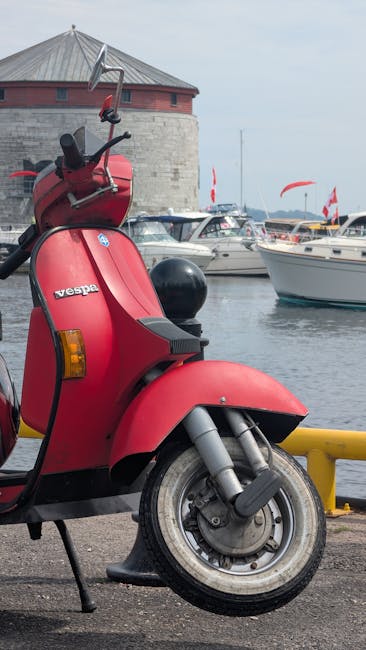 A close-up view of a red Vespa scooter parked on a paved surface near a waterfront, with boats moored in the background. The scooter is positioned slightly to the left, showing the front wheel, part of the seat, and the handlebar with rearview mirrors. Behind the scooter, a yellow safety barrier runs parallel to the water's edge. The scene includes a historic round stone building with a conical metal roof, situated on the opposite side of the marina, and several boats with white hulls and red flags docked alongside. The sky is overcast, casting diffused lighting over the area, and the water is calm, reflecting the boats and structures. This image captures elements relevant to home relocation logistics, illustrating the transportation environment near the property on Kingston Road to New Malden, where [COMPANY_NAME], specialising in removals and furniture transport, might coordinate the loading process of household belongings.