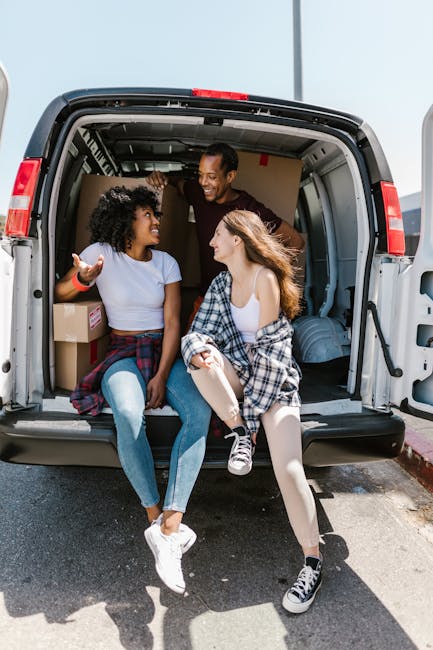 Two young women sitting inside the open back of a moving van during a home relocation process, engaged in conversation and smiling at each other. The van's interior contains cardboard boxes, some wrapped in plastic or paper, along with a few larger items possibly wrapped in moving blankets. One woman has curly black hair and is wearing a white t-shirt with a red wristband, while the other has long, wavy light brown hair and is dressed in a white top with a checkered shirt draped over her lap. The van is parked on a paved area, with sunlight illuminating the scene, indicating daytime. Visible features include the vehicle’s interior with cargo space, packaging materials, and the women’s casual clothing suggesting a friendly, organized packing and moving operation supported by [COMPANY_NAME], specialized in removals for house and furniture transport within the area covered by [PAGE_TITLE].