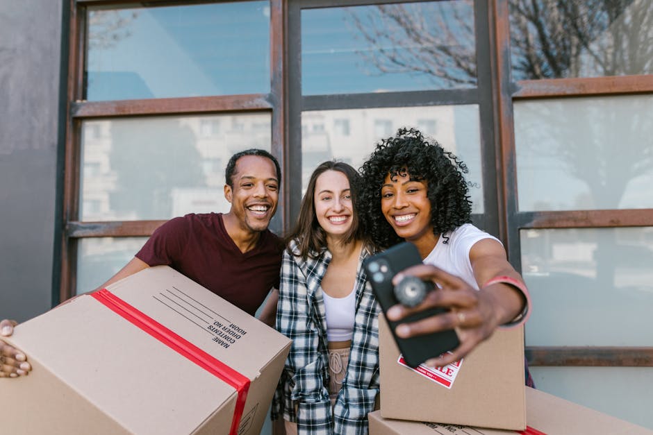 Three individuals standing outdoors in front of a large window with a metal frame, engaged in a home relocation or moving process. The person on the left holds a cardboard box with red packing straps labeled 'Moving Boxes,' while the person on the right is taking a selfie with a smartphone, holding another box. The middle individual smiles broadly, with a plaid shirt over casual attire. There are additional packing materials and moving boxes visible, indicating a packing phase of furniture transport and home moving. The background shows a partly blurred urban setting with trees and buildings, and the natural daylight illuminates the scene. The setting suggests a friendly, professional moving service environment that involves packing, loading, and transporting household items as part of a comprehensive removals process, aligned with services offered by manwithvanmotspurpark.co.uk.