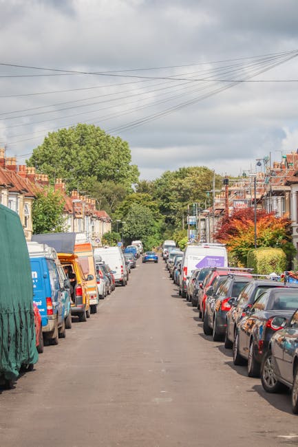 A residential street lined with closely spaced terraced houses featuring red brick facades and front gardens, with cars parked along both sides of the narrow, asphalt-paved road. The vehicles include small hatchbacks, vans, and larger commercial vehicles, some of which are covered with protective tarps or plastic wraps. Overhead, electrical wires stretch across the sky, which is partly cloudy. In the distance, a manwithvanmotspurpark.co.uk van is visible, indicating a home relocation or moving service. The street is surrounded by lush green trees and well-maintained shrubbery, suggesting a suburban neighbourhood. The scene captures the typical environment where furniture and household items are loaded onto vans during moving or packing and moving processes, reflecting the logistics involved in local house removals.