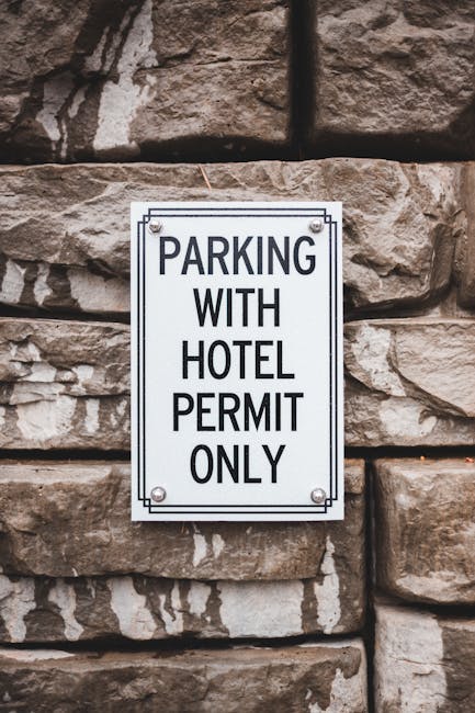 A metal sign affixed to a rough stone wall indicates parking restrictions, reading 'PARKING WITH HOTEL PERMIT ONLY.' The sign is mounted with four bolts, one in each corner, and is positioned at a height suitable for visibility. The surrounding stone wall appears weathered, with irregular-sized stones cemented together, providing a textured background. This image relates to house removals and moving services, such as those offered by manwithvanmotspurpark.co.uk, as it highlights parking permit requirements often relevant for efficient furniture transport and home relocation logistics within the Motspur Park area.
