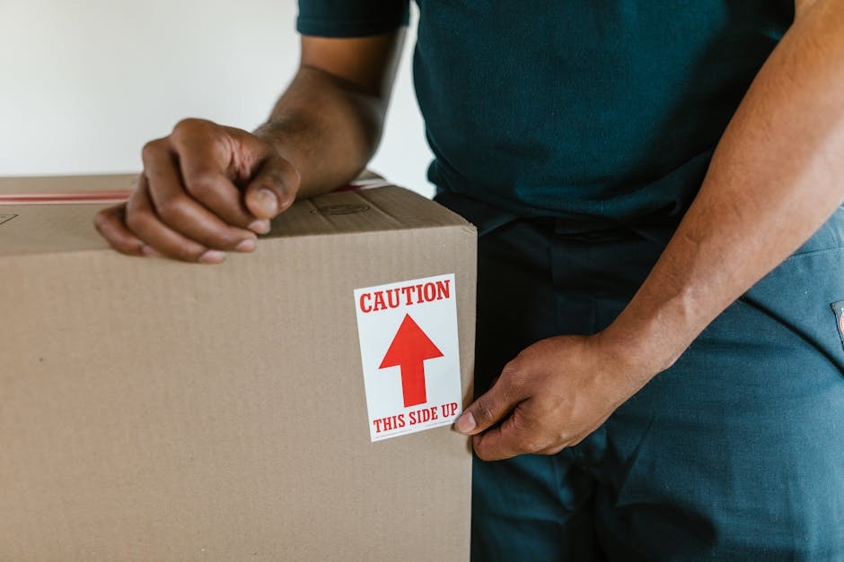 Close-up of a person wearing a dark green T-shirt and blue trousers, holding a large cardboard box secured with packing tape. The box has a red and white caution label indicating 'Caution: This Side Up' with an upward-pointing arrow. The individual is inside a room, possibly at a doorway or near a moving vehicle, preparing the box for transport during a house relocation. The background is plain and unobtrusive, emphasizing the focus on the packing process. This image illustrates packing and handling procedures typical of furniture transport and home moving services, such as those offered by manwithvanmotspurpark.co.uk, highlighting the importance of proper packaging during a same-day removal or urgent home move.