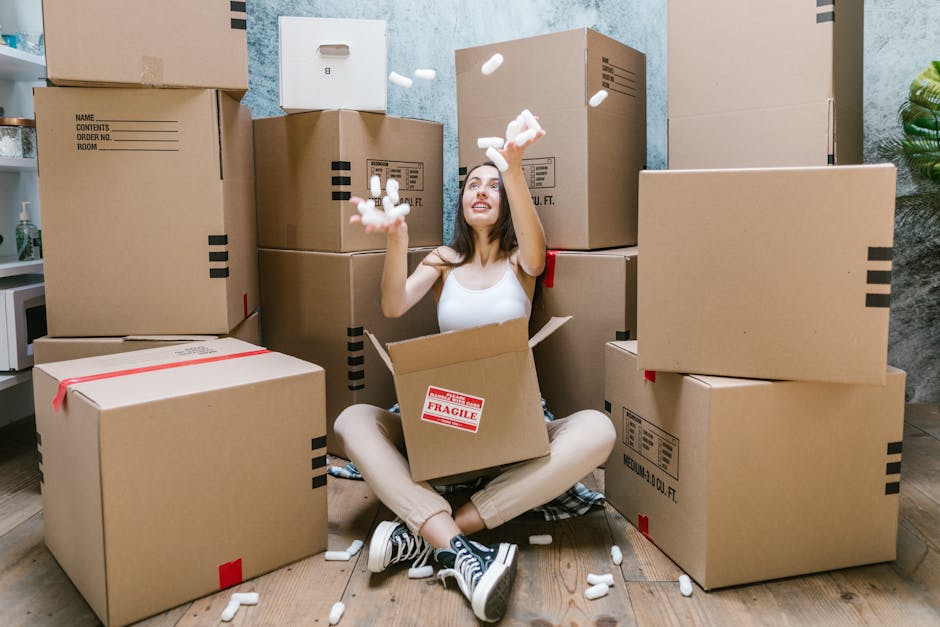 Close-up of a person wearing a dark green T-shirt and blue trousers, holding a large cardboard box secured with packing tape. The box has a red and white caution label indicating 'Caution: This Side Up' with an upward-pointing arrow. The individual is inside a room, possibly at a doorway or near a moving vehicle, preparing the box for transport during a house relocation. The background is plain and unobtrusive, emphasizing the focus on the packing process. This image illustrates packing and handling procedures typical of furniture transport and home moving services, such as those offered by manwithvanmotspurpark.co.uk, highlighting the importance of proper packaging during a same-day removal or urgent home move.