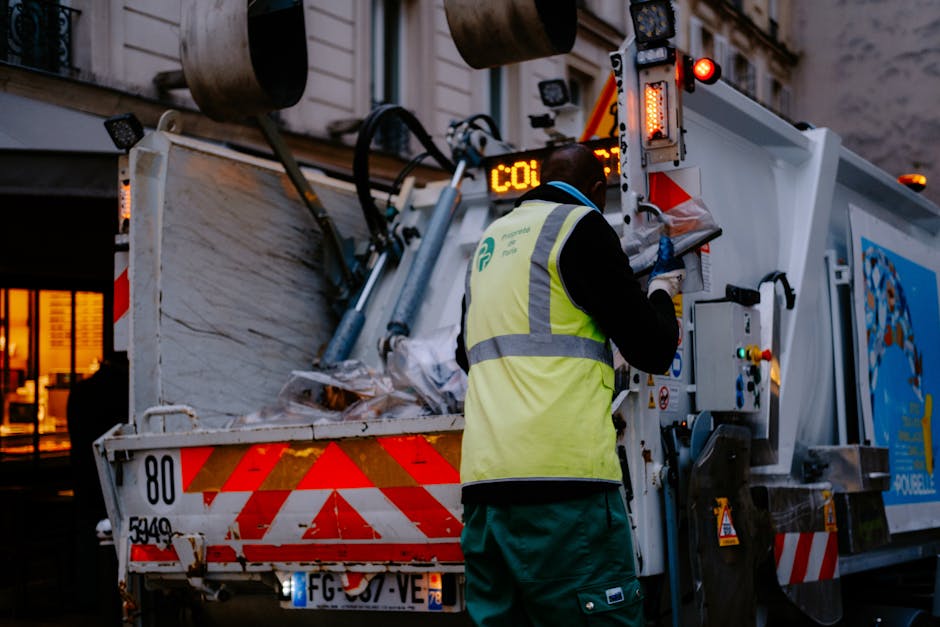 A worker wearing a high-visibility yellow vest is operating a waste collection vehicle with a white and red striped rear, situated on a paved area outside a building during dusk. The worker, with their back to the camera, is loading or unloading waste materials, which include cardboard boxes, plastic packaging, and fabric sheets, into the vehicle's rear compartment. The vehicle is equipped with loading and control equipment, with visible lights and electronic displays on the back. The interior of the waste truck shows various mechanical components, hoses, and safety features. Nearby, there are nearby windows of a building with warm lighting, indicating an urban environment. This scene exemplifies waste disposal services associated with home relocation or furniture transport, as handled by companies like manwithvanmotspurpark.co.uk, supporting efficient moving logistics and packing processes.