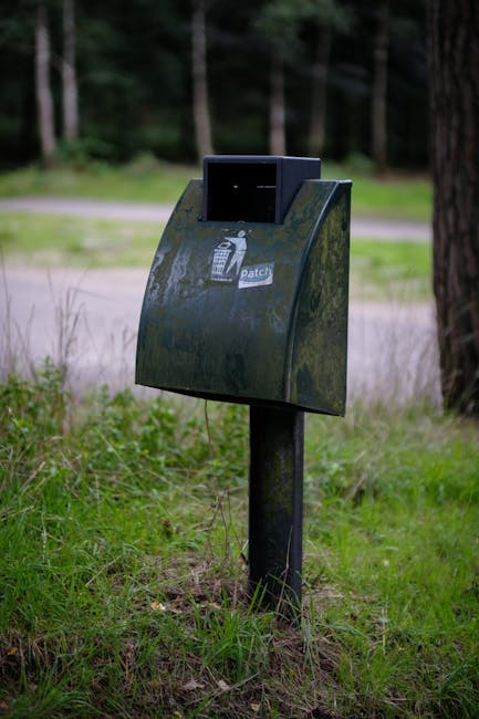 A black communal waste bin with a hinged lid, mounted on a metal post situated on a grassy area beside a paved path. The front of the bin features a small sticker with a white trash can icon and the word 'patch', suggesting it is part of a local waste collection scheme. The surrounding environment includes tall trees and green grass, indicating a park-like setting. The bin is positioned near a wooden fence and appears weathered with some moss or dirt visible on its surface. In the background, out-of-focus natural features such as trees and the paved pathway are visible, contributing to an outdoor, public space scene. This image reflects waste disposal infrastructure that might be found in outdoor or residential communal areas, relevant to home and business waste management in the context of relocation or removal services by companies like manwithvanmotspurpark.co.uk.