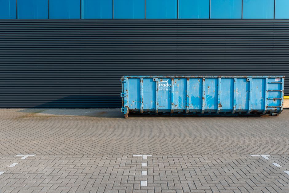 A worker wearing a high-visibility yellow vest is operating a waste collection vehicle with a white and red striped rear, situated on a paved area outside a building during dusk. The worker, with their back to the camera, is loading or unloading waste materials, which include cardboard boxes, plastic packaging, and fabric sheets, into the vehicle's rear compartment. The vehicle is equipped with loading and control equipment, with visible lights and electronic displays on the back. The interior of the waste truck shows various mechanical components, hoses, and safety features. Nearby, there are nearby windows of a building with warm lighting, indicating an urban environment. This scene exemplifies waste disposal services associated with home relocation or furniture transport, as handled by companies like manwithvanmotspurpark.co.uk, supporting efficient moving logistics and packing processes.