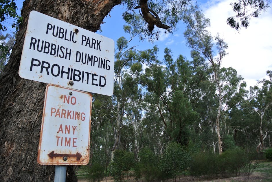 A close-up view of two roadside signs attached to a large tree trunk in a park. The upper sign is white with black text stating 'PUBLIC PARK RUBBISH DUMPING PROHIBITED', indicating that dumping rubbis